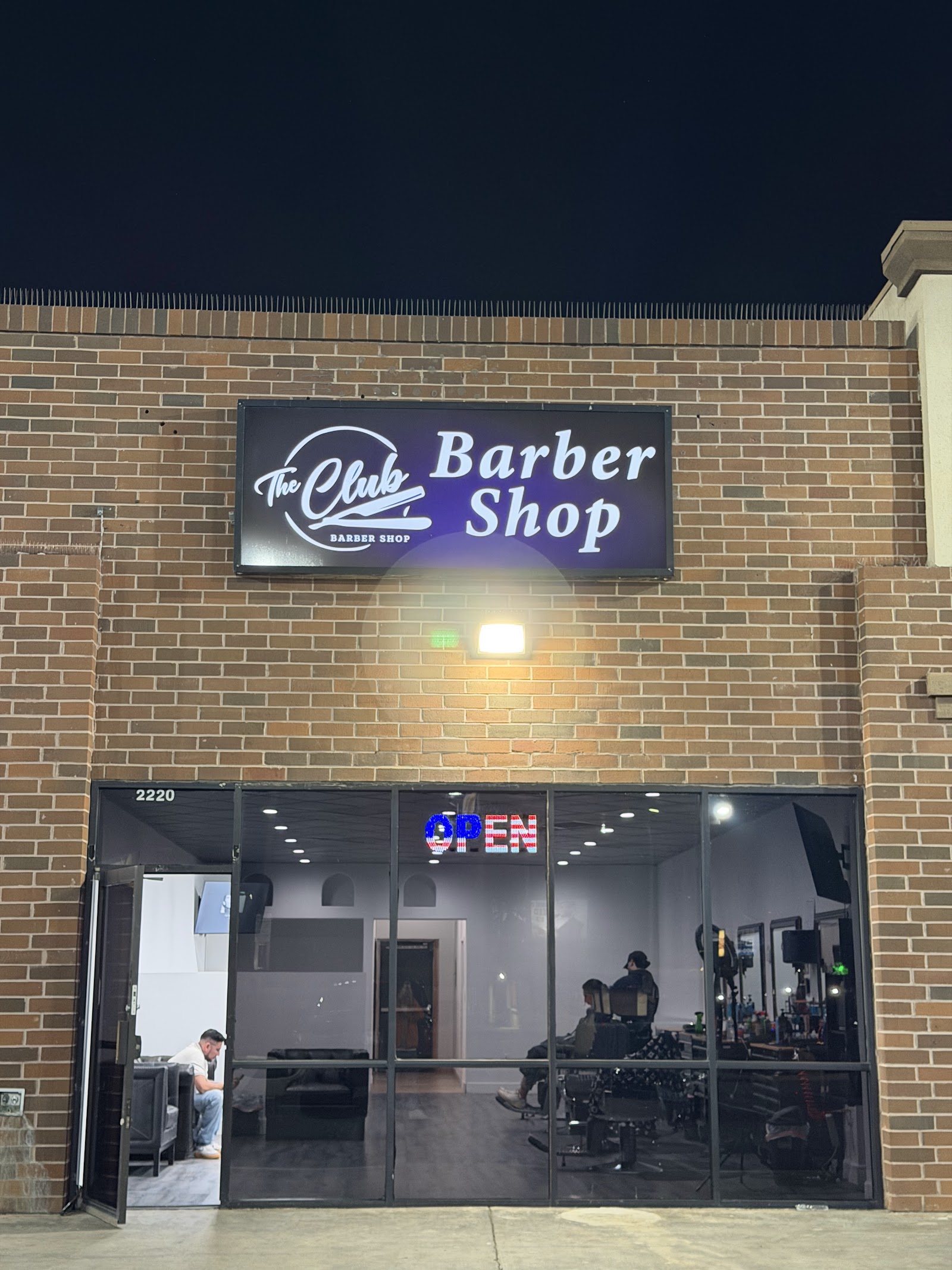 The Club Barber Shop illuminated storefront sign at night with an OPEN sign and brick facade