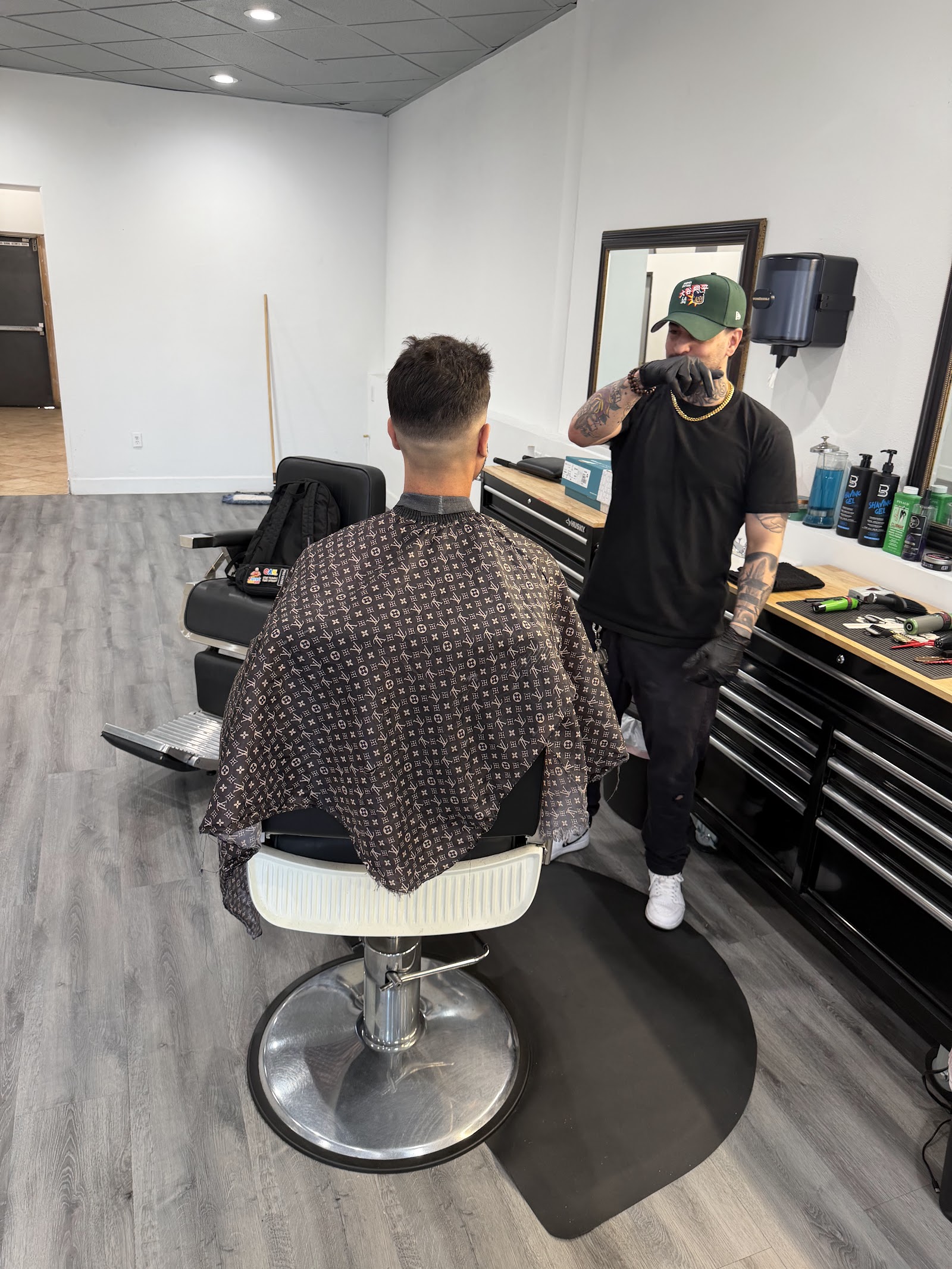 Barber in a green cap standing near a workstation while a client sits in a chair with a patterned cape
