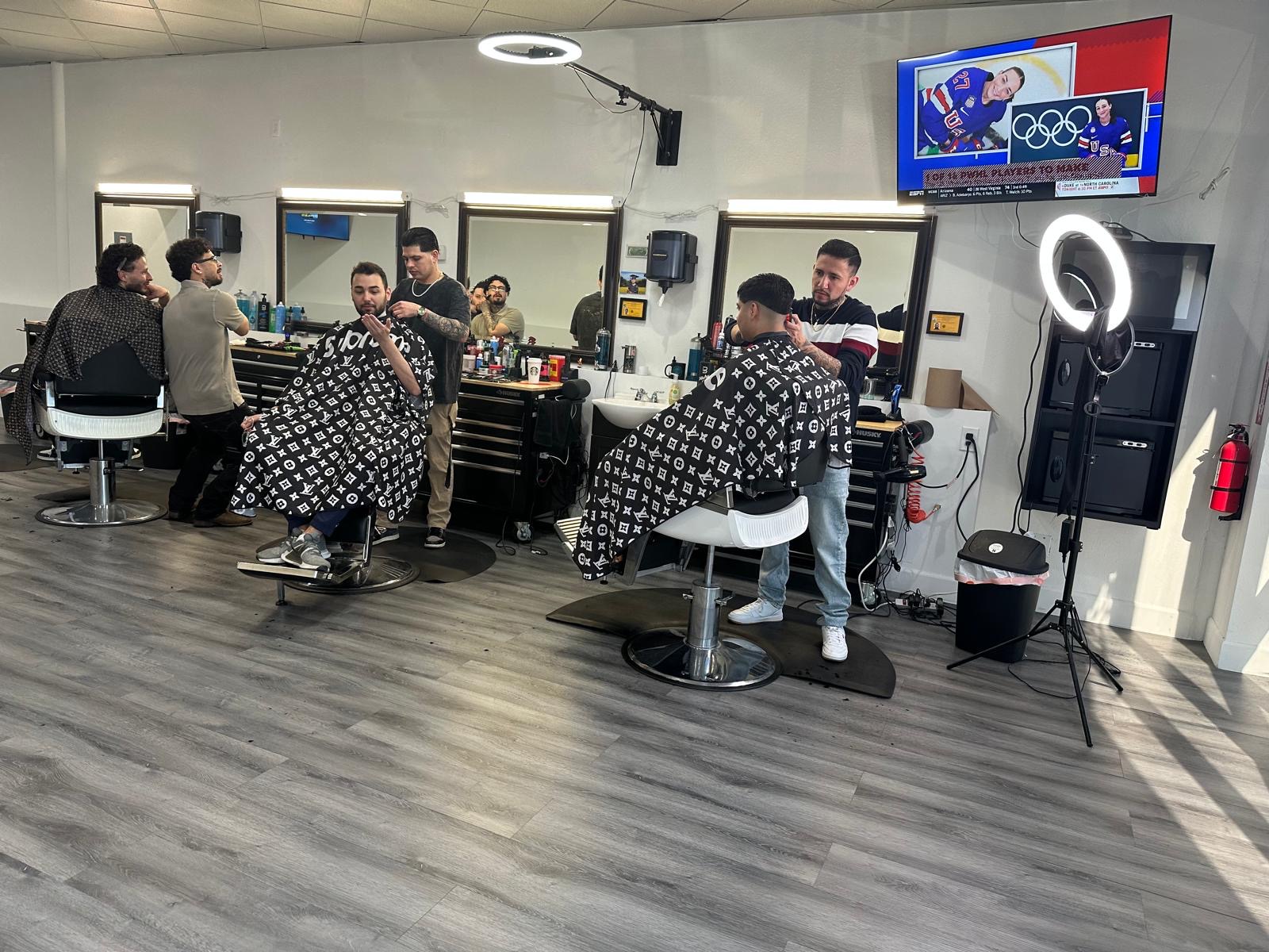 Busy barbershop with multiple barbers cutting clients' hair, sports on the TV and ring lights overhead